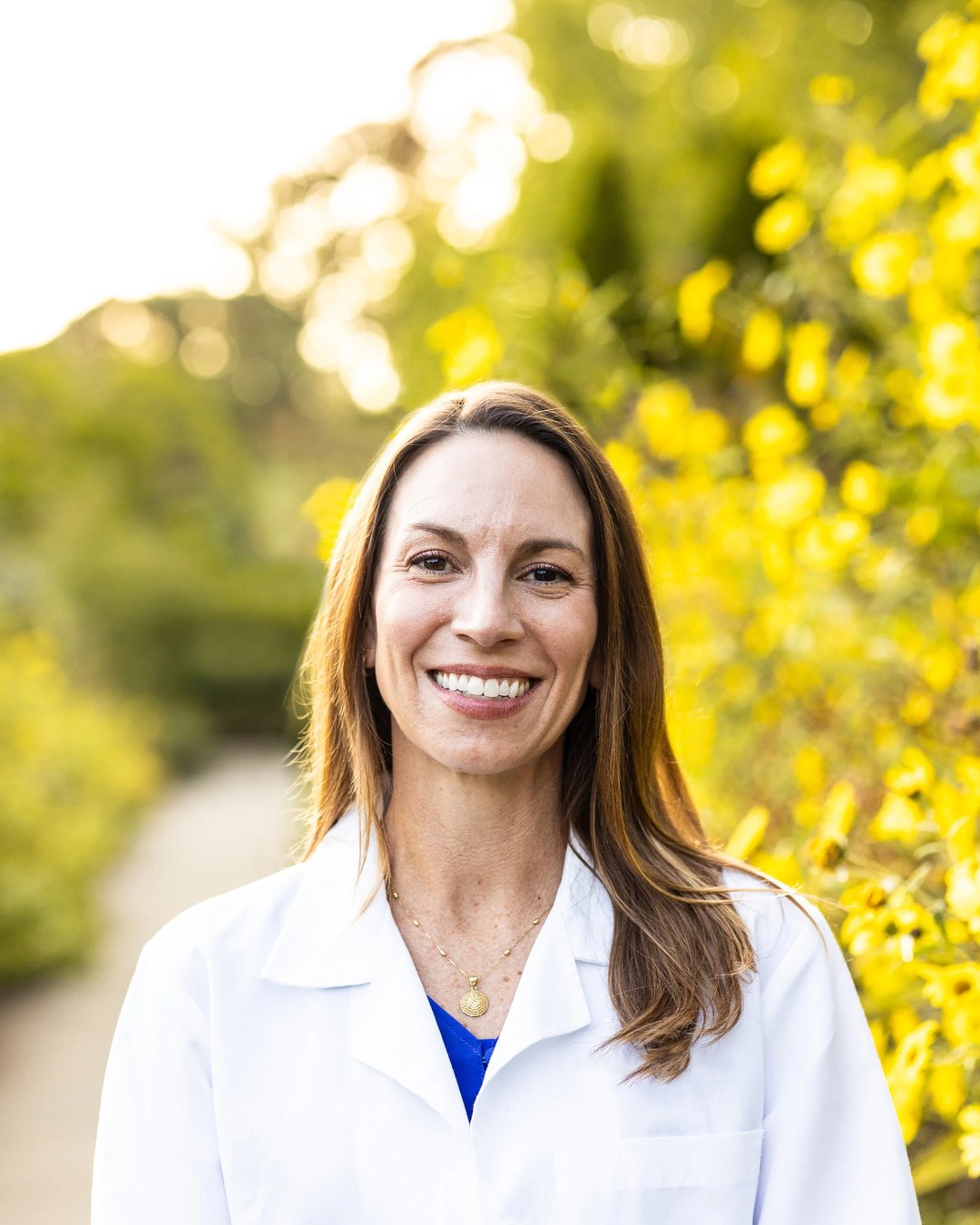 Dr. Cara Jacob in a white coat, smiling on a garden path beside yellow flowers
