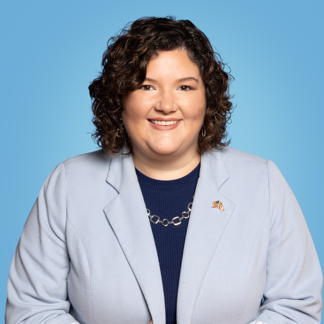 Jessica Miranda in a light blue blazer smiling in a professional portrait on a sky blue background