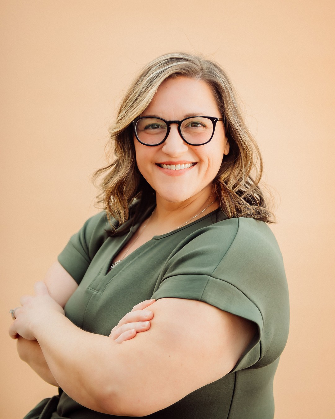 Jordan Haire smiling with arms crossed, wearing glasses and an olive top, against a peach backdrop