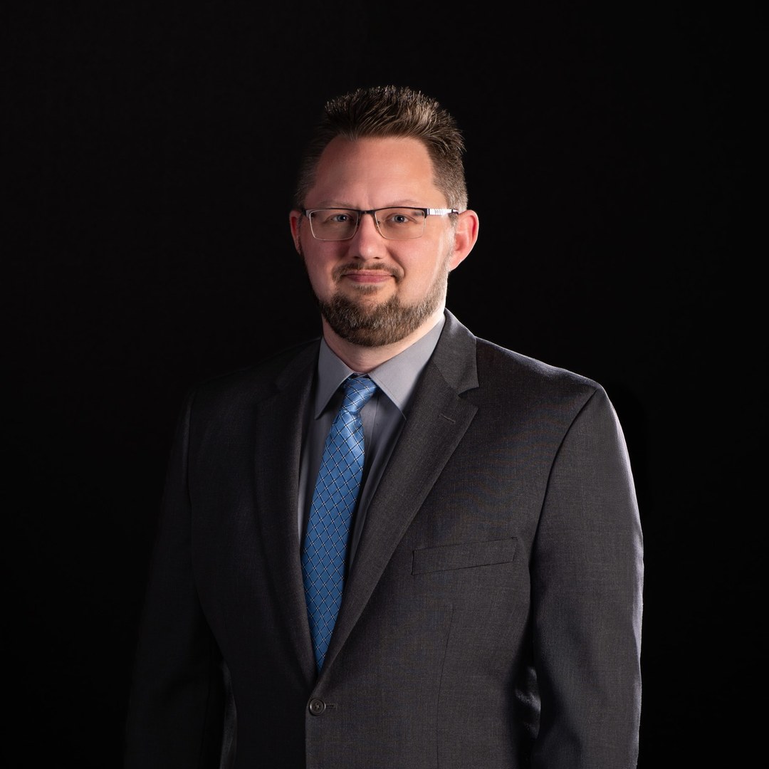 Mike Baker in a gray suit and blue tie with glasses and a beard, smiling in a professional portrait on a dark background