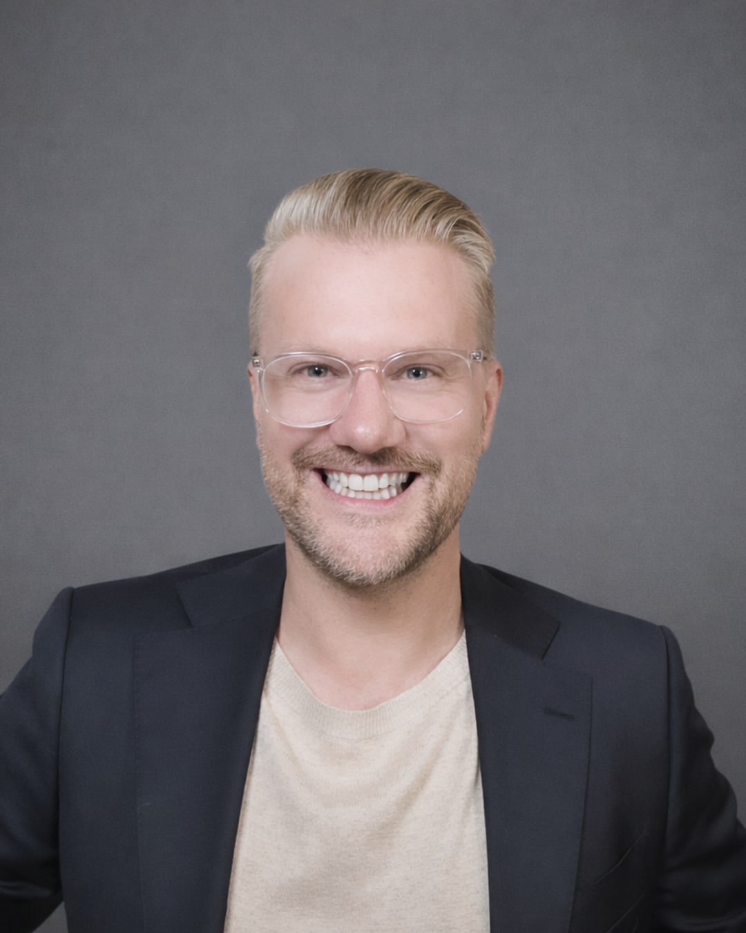 Paul Kurtz smiling, wearing clear-framed glasses and a navy blazer over a cream tee, against a gray studio backdrop